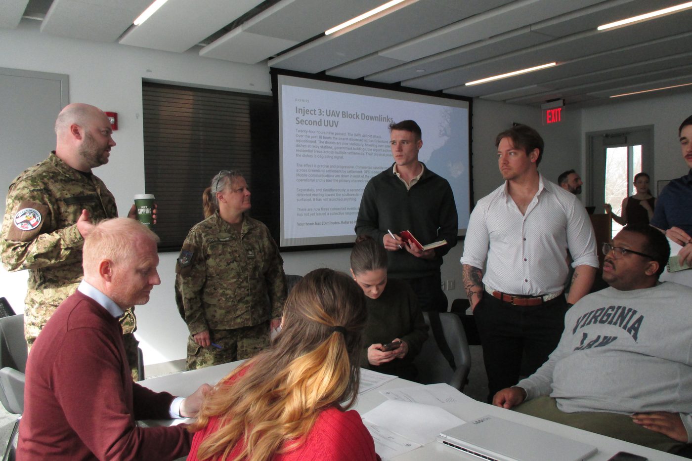 Participants and allied officers around a simulation table, a scenario inject on screen behind