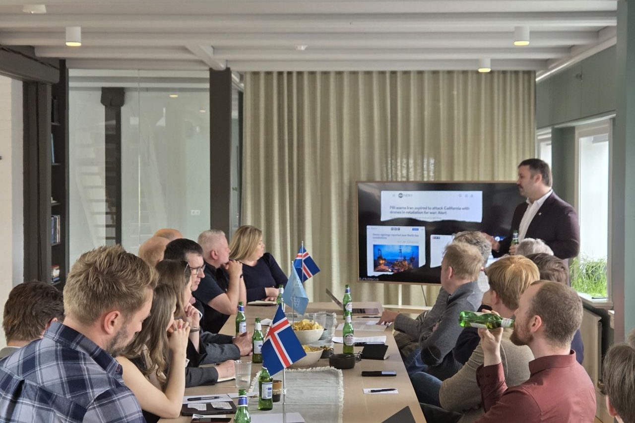 A long table of participants in Reykjavik, Icelandic flags on the table, drone incident coverage on screen